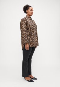 Chemise à imprimé léopard avec des manches longues, fentes sur les côtés ; associée à un pantalon noir ajusté et des escarpins à bout pointu avec un accent métallique.