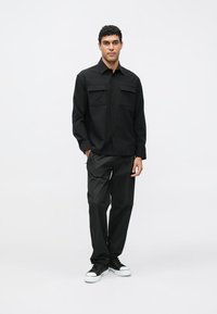 Mustast nööpidega särk kahe rinnataskuga, kombineeritud mustade pükstega. Modell kannab musti tenniseid. Sile kangas, lihtne disain, lõdvestunud istuvus.