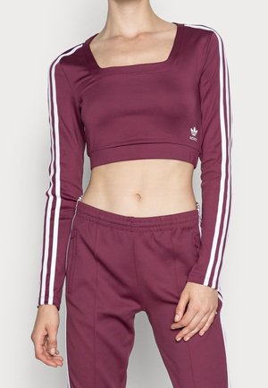 Mujer con crop top granate de Adidas y pantalones deportivos a juego con rayas blancas en las mangas y las perneras.