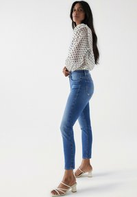 Jean skinny en denim bleu avec une taille haute, associé à un blouse blanche à pois et des sandales blanches à talons blocs avec des lanières.