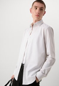 Camisa blanca de manga larga con cuello clásico, textura suave y rayas tonales. Lleva pantalones negros y un bolso negro.