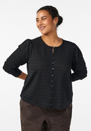 Usmievavá žena s vlasmi zviazanými do drdola má na sebe čiernu textúrovanú blúzku s gombíkmi a naberanými rukávmi a hnedé nohavice, ruky za chrbtom.