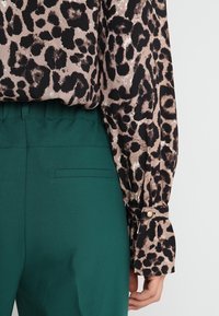 Leopardenmuster-Bluse in Beige und Schwarz mit Knopfmanschetten. Kombiniert mit grünen, hoch taillierten Hosen mit einer dezenten Gesäßtasche.