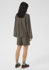 Kaki garām piedurknēm tops ar brīvu piegulšanu un kroku detaļām, saskaņots ar atbilstošām šortiem. Modelis valkā teksturētas pelēkas zeķes un brūnus apavus.