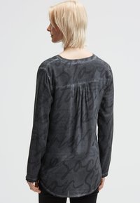 Blonde Person, die ein langärmliges, dunkelgraues Shirt mit großen, verblassten gedruckten Text trägt, mit dem Rücken zu einem hellen Hintergrund gewandt.