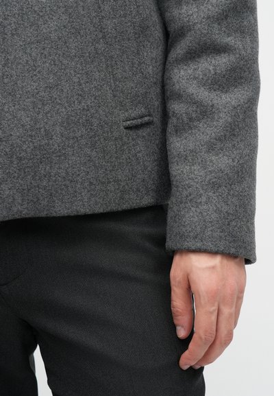 Šedý vlněný blazer s hladkou texturou, klopovými reverzy a detailem jednoho kapsy, spárovaný s tmavými texturovanými kalhotami a rukou odpočívající na boku.