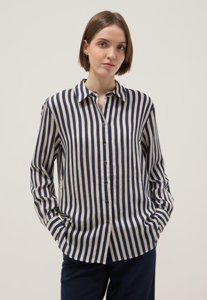 Mujer con cabello corto y castaño, vistiendo una camisa abotonada de rayas verticales en azul marino y blanco, con las manos en los bolsillos, sobre un fondo liso.