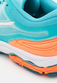 Zapatilla deportiva azul claro con parte superior de malla, acentos naranjas, amortiguación blanca, suela texturizada y elementos de diseño ondulados en el lateral.