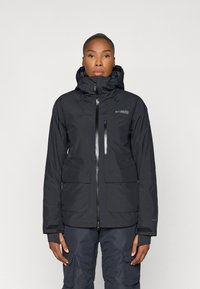 Chaqueta impermeable negra con capucha, cierre de cremallera frontal y múltiples bolsillos. Textura suave con un sutil logo en el pecho y los puños.