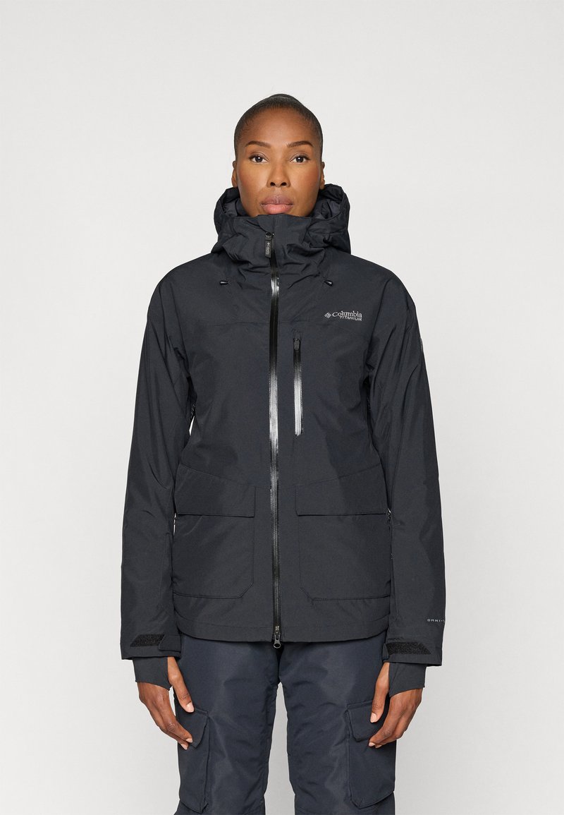 Chaqueta impermeable negra con capucha, cierre de cremallera frontal y múltiples bolsillos. Textura suave con un sutil logo en el pecho y los puños.