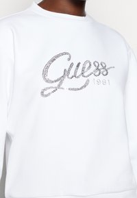 Felpa bianca con scollo tondo, decorata con un logo "Guess" in strass argentati e il testo "1981" in un font elegante.