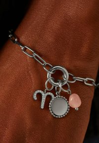 Zilveren kettingarmband met een charm in de vorm van een astrologisch symbool, een ronde spiegelcharme en een rozenkwarts accent, uitgestald op een achtergrond van textuur van huid.