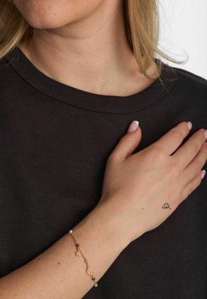 Arany karkötő színes gyöngyökkel és csillag díszítéssel, csuklón viselve. A póló sötét színű, kerek nyakú. A bőrszín világos.