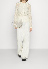 Häkelgestrickte cremefarbene Strickjacke mit Knopfverschluss und langen Ärmeln, kombiniert mit gemusterten weißen weit geschnittenen Hosen und einer hellgrauen Umhängetasche.
