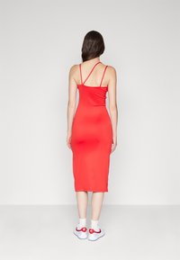 Robe midi rouge ajustée avec de fines bretelles spaghetti, texture lisse et silhouette épurée. Associée à des baskets blanches avec des accents rouges.