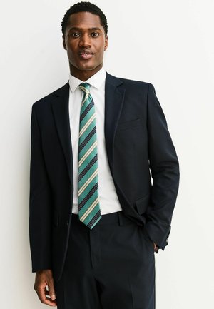 Bărbat în costum închis la culoare, cu cămașă albă și cravată în dungi verde teal, bej și negru, stând pe un fundal alb simplu, cu o mână în buzunar.