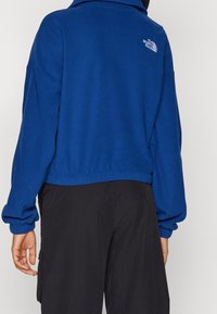 Niebieski polarowy sweter z wysokim kołnierzem i elastycznymi mankietami. Posiada białe haftowane logo na górnej części pleców. Noszony z czarnymi spodniami.