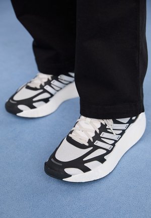 Persoon draagt zwart-witte sportschoenen met een witte zool, staand op een blauwe ondergrond, benen zichtbaar van de kuit naar beneden.