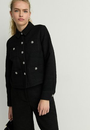 Black textured jacket featuring a collared design, four front pockets, and silver button accents. Styled with matching black pants.
