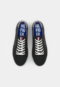 Zapatillas negras con cordones blancos, acentos grises y una plantilla azul con "TOMMY HILFIGER". Diseño plano y de punta redonda con una textura suave.