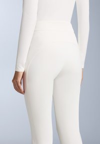 Fehér, magas derékú, testhez simuló leggings, sima textúrával és minimális varrási részletekkel. Hátulról látható, egy kéz a csípőn pihen.