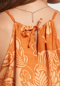 Back view of a woman wearing an orange floral sleeveless dress with a tied bow at the upper back and a gold chain necklace.