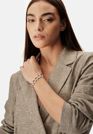 Bracciale a catena in argento indossato al polso, con maglie curve intrecciate. Il modello indossa un blazer beige testurizzato.
