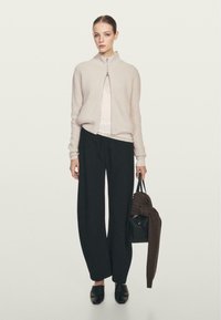 Maglione zip-up a coste grigio chiaro, top bianco, pantaloni neri a gamba larga, mocassini neri e una borsa nera con una sciarpa lavorata a maglia marrone.