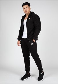 Zwarte zip-up hoodie met een wit logo, gecombineerd met zwarte sweatpants en zwarte sneakers. De outfit heeft een soepele textuur en een sportieve pasvorm.