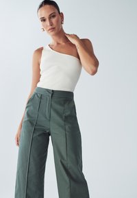 Biely rebrovaný top na jedno rameno v kombinácii s vysokými zelenými nohavicami s širokými nohavicami. Nohavice majú záhyby a hladký povrch. Zlatozlaté náušnice dopĺňajú vzhľad.