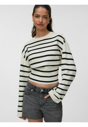 Ung kvinde iklædt en hvid og sort stribet cropped sweater med lange ærmer og mørkegrå denimshorts, poserer med den ene hånd i lommen.