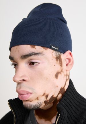Granatowa dzianinowa czapka z prążkowanego splotu z podwiniętym mankietem, z czarną metką z białym napisem. Model ma na sobie teksturowany czarny sweter na zamek.
