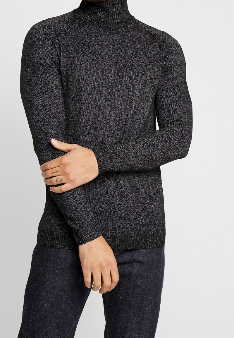 Pull à col roulé noir avec un design en maille texturée, manches longues et poignets côtelés, porté avec un pantalon en denim foncé.