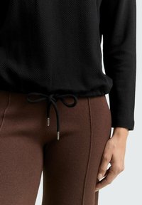 Schwarzes, strukturiertes Oberteil mit einem Kordelzug an der Taille und silbernen Ösen, kombiniert mit braunen, enganliegenden Hosen mit seitlicher Nahtdetail.