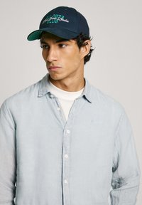 Blue linen button-up shirt with a collar, paired with a dark blue baseball cap featuring green embroidery. Textured fabric, casual fit.