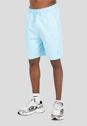 Uomo che indossa pantaloncini azzurri con coulisse, calzini bianchi Nike e scarpe da ginnastica bianche con dettagli neri, in piedi davanti a uno sfondo semplice.