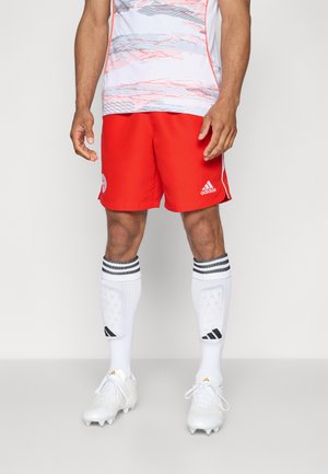 Shorts de football rouges avec des accents blancs, associés à des chaussettes blanches arborant des rayures noires, et des crampons blancs, portés par une silhouette debout.