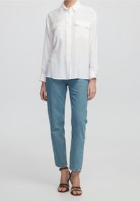 Chemise blanche à boutons avec deux poches poitrine, associée à un jean bleu clair et des talons striés marron. Tissu lisse, coupe décontractée, design simple.