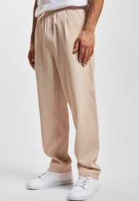 Pantalones de tono beige con un ajuste holgado y frente con pliegues, que cuentan con una cinturilla elástica. Combinados con zapatillas blancas.