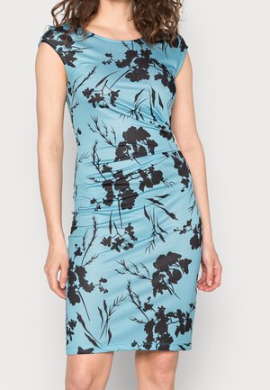 Vrouw draagt een nauwsluitende, knie-lange blauwe jurk met zwarte bloemen- en takkenpatronen, korte mouwen en lichte plooien bij de taille.