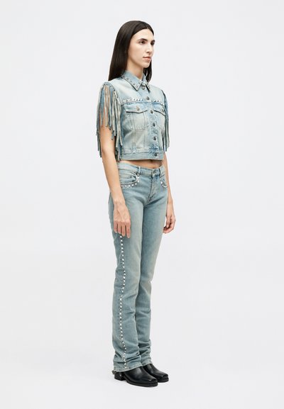Γυναίκα που στέκεται φορώντας ανοιχτόγαλανό denim γιλέκο με φράντζες στα μανίκια και τζιν παντελόνι με καρφιά στο ίδιο χρώμα, φοράει μαύρες μπότες μπροστά σε απλό φόντο.