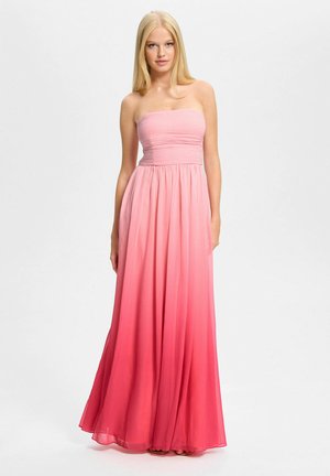 Donna bionda che indossa un vestito lungo fino al pavimento senza spalline con sfumatura di colore dal rosa al rosso, in piedi davanti a uno sfondo bianco.