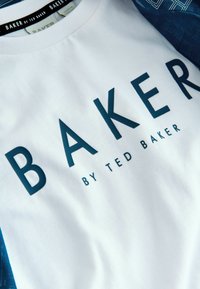 Biała koszula z dużym granatowym napisem "BAKER" oraz mniejszym napisem "BY TED BAKER" nadrukowanym poniżej metek na ciemnoniebieskim ubraniu.