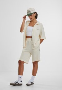 Krémový súprava s ryhovaným vzorom, ktorá obsahuje košeľu s krátkym rukávom a zodpovedajúce šortky. Pod ňou je biela crop top a béžová čiapka so zeleným okrajom.