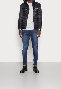 Zwarte pufferjas met capuchon, grijze sweatshirt, blauwe skinny jeans en zwarte enkellaarsjes. De jas heeft een glanzende afwerking en een doorgestikt ontwerp.