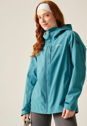 Femme portant une veste d'extérieur bleue à capuche avec un bandeau blanc, tenant une sangle grise, debout devant un fond beige.