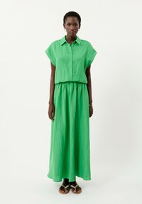Green linen maxi dress with short sleeves, button-down front, and elastic waistband. Model wears brown sandals with a slight heel.