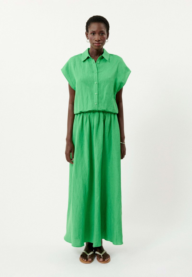 Green linen maxi dress with short sleeves, button-down front, and elastic waistband. Model wears brown sandals with a slight heel.