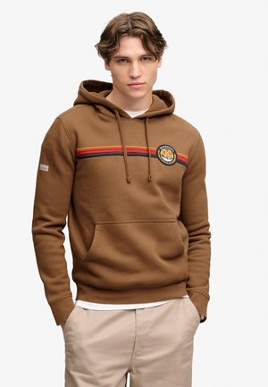 Ung mand iført en brun hættetrøje med røde, orange og sorte striber og et rundt mærke, hænder i beige bukselommer.