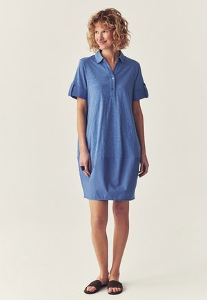 Frau mit lockigem Haar, die ein blaues Polokleid mit kurzen Ärmeln und schwarzen Slide-Sandalen trägt, steht und schaut lächelnd zur Seite.